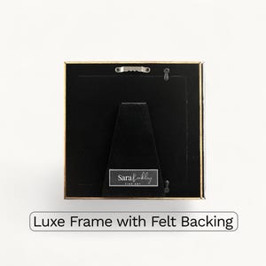 May include: Square picture frame with a gold border and black backing. The frame has a felt backing and a small metal hanger. The text "Luxe Frame with Felt Backing" is at the bottom.