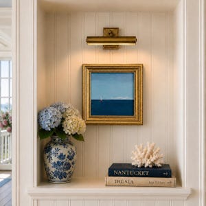 May include: A square painting in a gold frame depicting a blue sea and sky, hung below a brass picture light. Beside it sits a blue and white floral ceramic vase with hydrangeas, a white coral sculpture, and two books titled NANTUCKET and THE SEA.