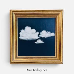 May include: A square painting featuring three white clouds against a dark navy blue background, enclosed in a decorative gold frame. The text Sara Beckley Art is printed in black below the frame. The artwork displays a clear sky scene.