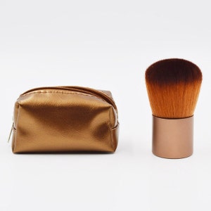 May include: A gold-colored makeup brush with a brown and orange bristle head, and a matching gold-colored zippered pouch.