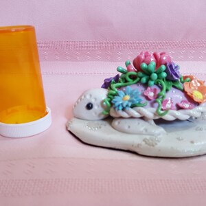 Pu&ograve; includere: Una statuetta di tartaruga in ceramica bianca con un guscio rosa decorato con fiori colorati e foglie verdi. La tartaruga &egrave; seduta su una base bianca.