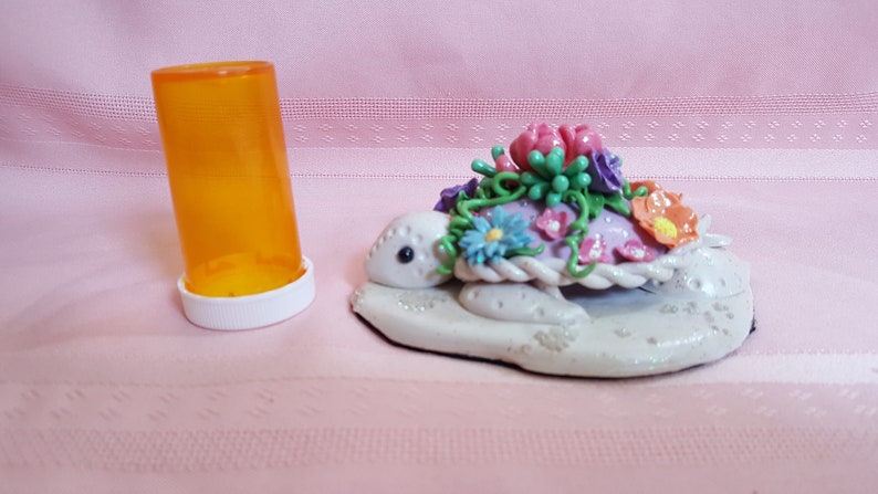 Pu&ograve; includere: Una statuetta di tartaruga in ceramica bianca con un guscio rosa decorato con fiori colorati e foglie verdi. La tartaruga &egrave; seduta su una base bianca.