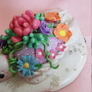 Pu&ograve; includere: Una statuetta in ceramica bianca con una base viola e una variet&agrave; di fiori e vegetazione colorati in cima. I fiori sono fatti di pasta polimerica e hanno una finitura scintillante.