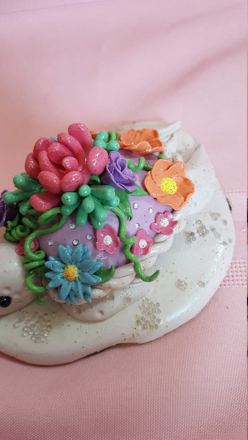 Pu&ograve; includere: Una statuetta in ceramica bianca con una base viola e una variet&agrave; di fiori e vegetazione colorati in cima. I fiori sono fatti di pasta polimerica e hanno una finitura scintillante.
