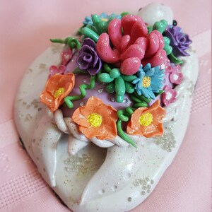 Pu&ograve; includere: Una statuetta di tartaruga in ceramica bianca con un arrangiamento floreale rosa, verde e arancione sul dorso. La tartaruga &egrave; ricoperta di glitter e ha una base viola.