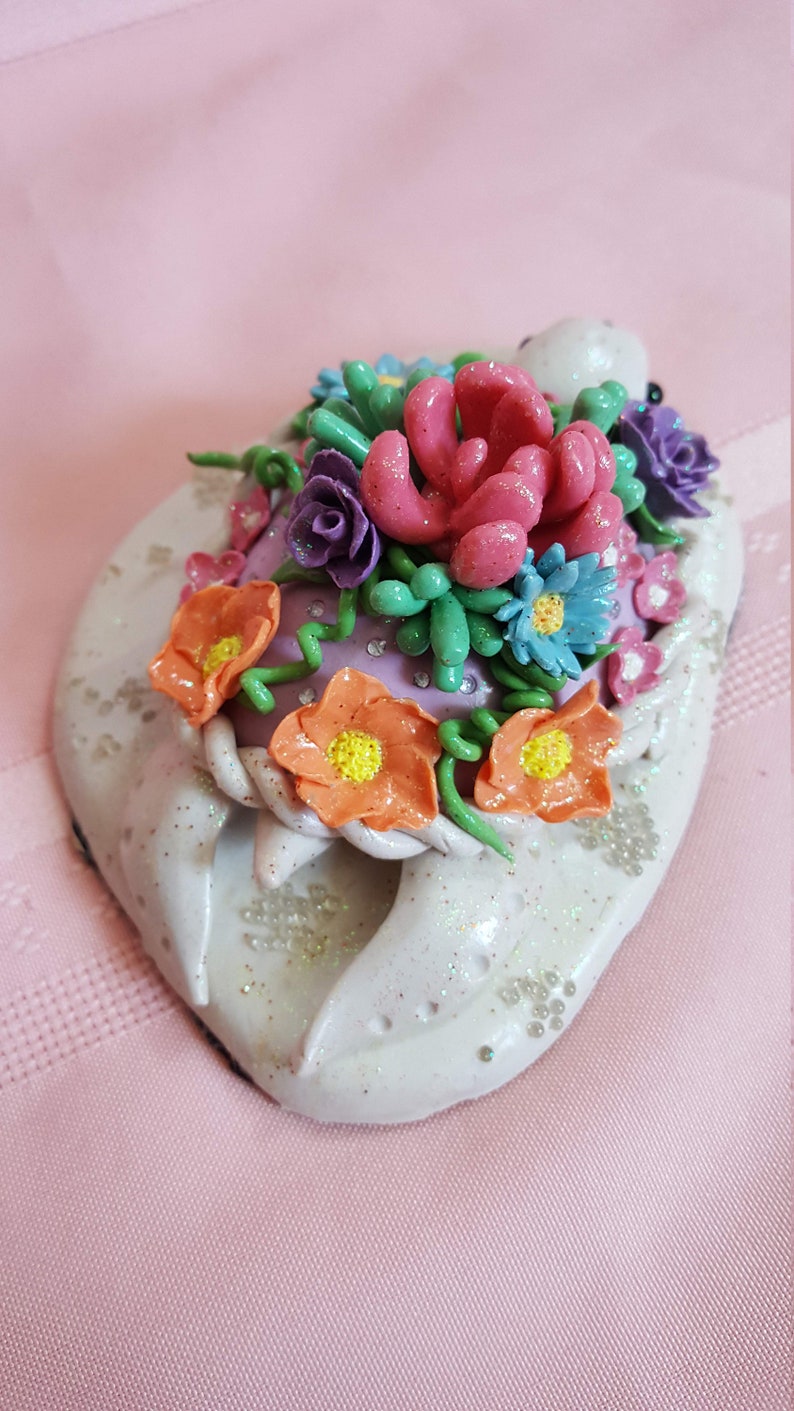 Pu&ograve; includere: Una statuetta di tartaruga in ceramica bianca con un arrangiamento floreale rosa, verde e arancione sul dorso. La tartaruga &egrave; ricoperta di glitter e ha una base viola.