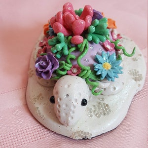 Pu&ograve; includere: Una statuetta di tartaruga in ceramica bianca con un guscio viola decorato con viti verdi, fiori rosa, viola e blu e strass. La tartaruga &egrave; seduta su una base bianca con glitter bianco.