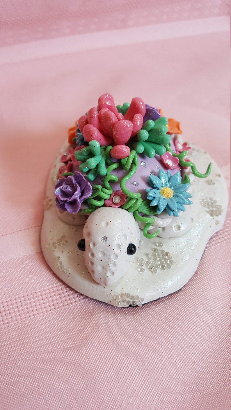 Pu&ograve; includere: Una statuetta di tartaruga in ceramica bianca con un guscio viola decorato con viti verdi, fiori rosa, viola e blu e strass. La tartaruga &egrave; seduta su una base bianca con glitter bianco.