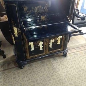 May include: Black antique cabinet with gold floral and figure accents. The cabinet has a drop-down front and two doors. The cabinet is on short, curved legs. The interior has shelves and compartments. The cabinet is likely made of wood.