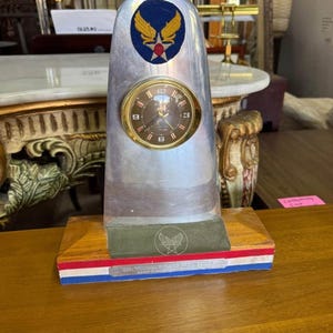 May include: A vintage desk clock with an aluminum body and a wooden base. The clock face is gold-toned with black numerals. The base features red, white, and blue stripes and an emblem. A blue and gold Air Force emblem is on the clock.