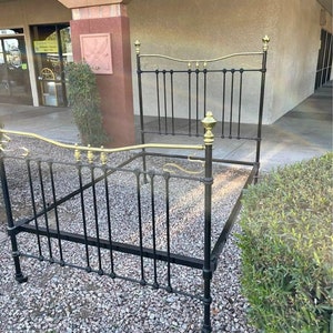 May include: Black metal bed frame with gold accents. The frame has a headboard with a curved top and a footboard with a straight top. The frame is sitting on a gravel surface.