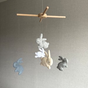 May include: A wooden baby mobile with five felt bunny toys. The bunnies are in shades of gray, white, and cream. The mobile is hanging from a wooden crossbar.