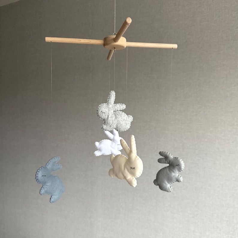 May include: A wooden baby mobile with five felt bunny toys. The bunnies are in shades of gray, white, and cream. The mobile is hanging from a wooden crossbar.