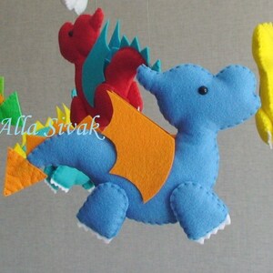 May include: A colorful felt mobile with four hanging figures: a blue dragon with orange wings, a red dragon with teal wings, a yellow dragon, and an orange dragon with green accents.