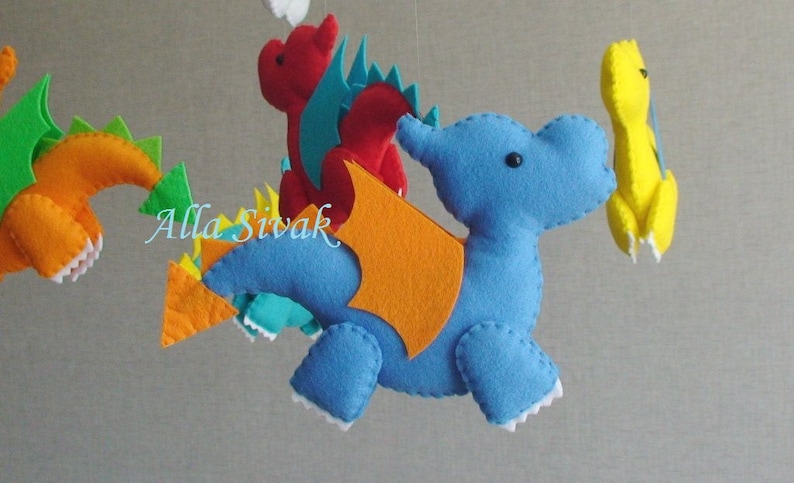 May include: A colorful felt mobile with four hanging figures: a blue dragon with orange wings, a red dragon with teal wings, a yellow dragon, and an orange dragon with green accents.