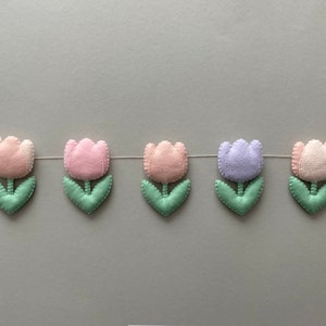 May include: A garland of felt tulips in pink, lavender, and light green. The tulips are strung together with white thread and have a simple, handmade look.