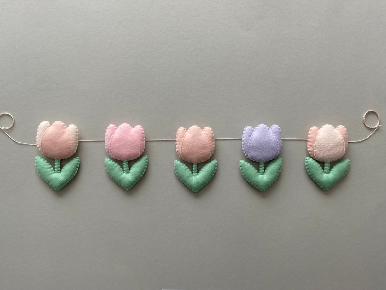 May include: A garland of felt tulips in pink, lavender, and light green. The tulips are strung together with white thread and have a simple, handmade look.