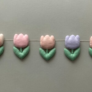 May include: A garland of felt tulips in pink, light pink, lavender, and white with green leaves. The garland is strung on a white thread.