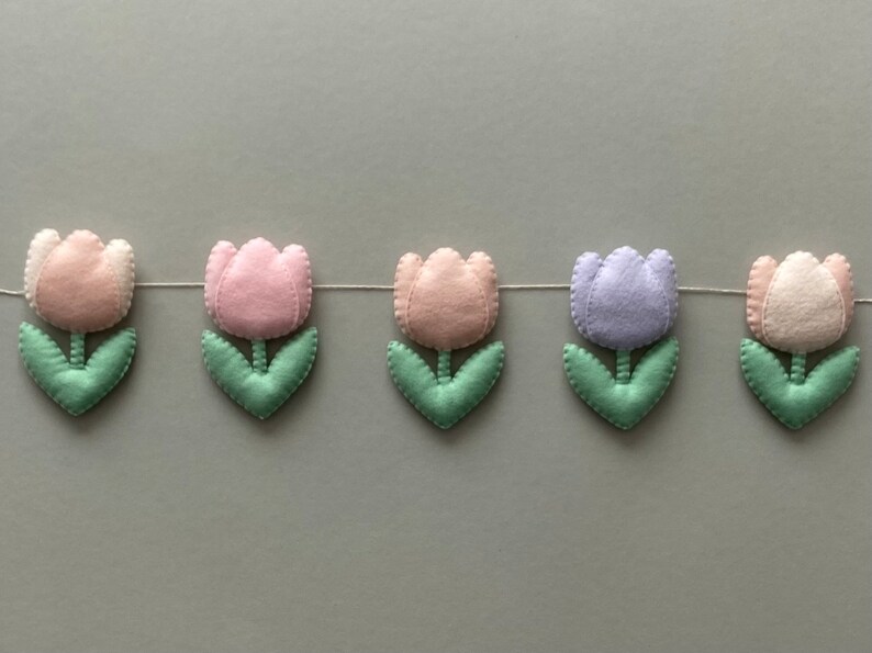 May include: A garland of felt tulips in pink, light pink, lavender, and white with green leaves. The garland is strung on a white thread.
