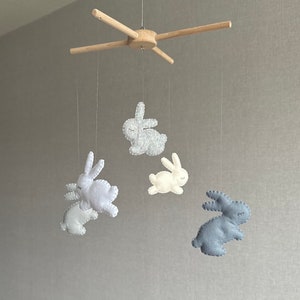 May include: A wooden mobile with four felt bunny toys hanging from it. The bunnies are in shades of gray, white, and blue.