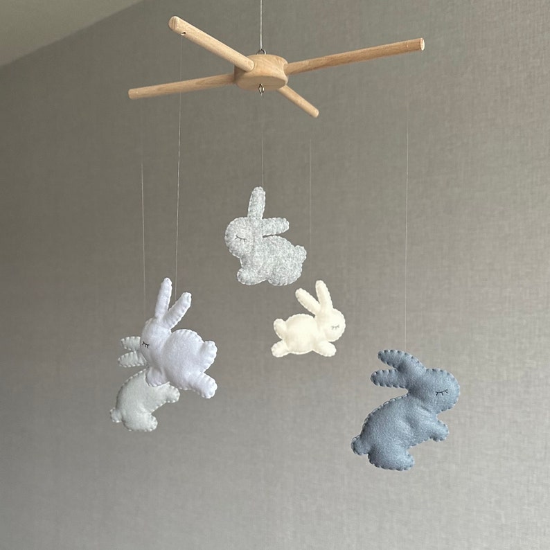 May include: A wooden mobile with four felt bunny toys hanging from it. The bunnies are in shades of gray, white, and blue.