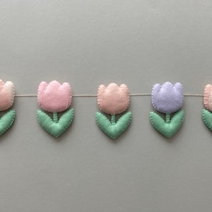 May include: A garland of felt tulips in pink, light pink, lavender, and white with green leaves. The garland is strung on a white thread.
