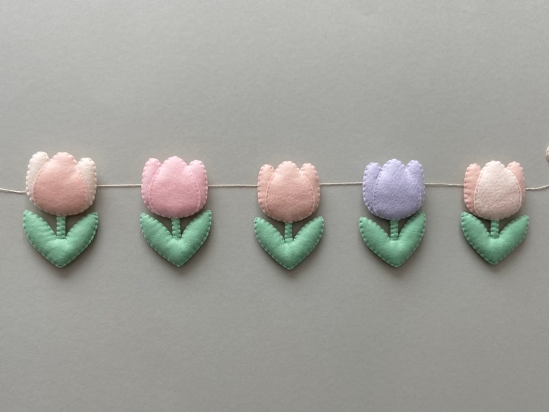 May include: A garland of felt tulips in pink, light pink, lavender, and white with green leaves. The garland is strung on a white thread.