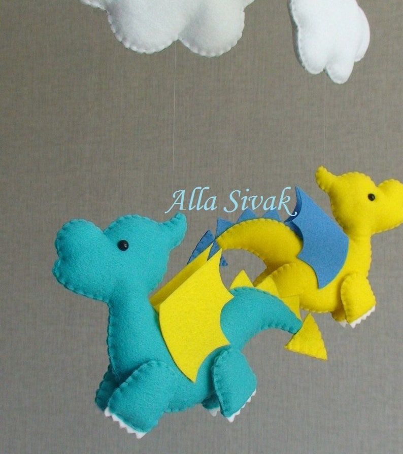 May include: A baby mobile with two felt dragons, one blue and one yellow, with white clouds hanging above. The dragons have yellow wings and blue accents.
