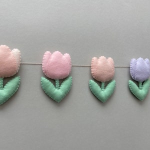 May include: A garland of felt tulips in pink, light purple, and white with green leaves. The tulips are strung together with white thread.