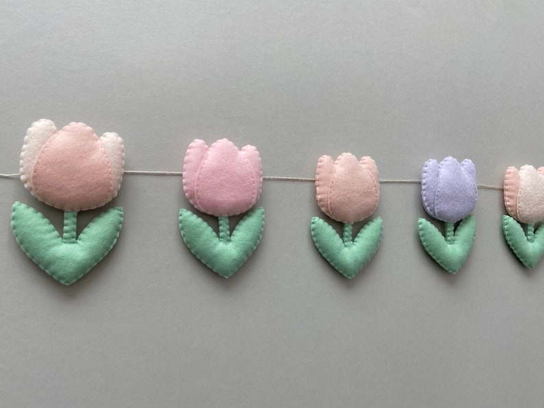 May include: A garland of felt tulips in pink, light purple, and white with green leaves. The tulips are strung together with white thread.