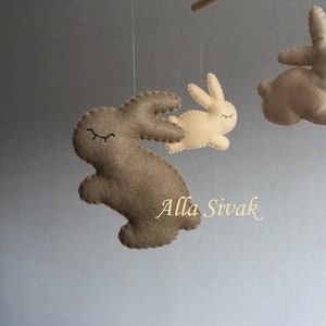 May include: A mobile with three felt bunny toys hanging from strings. The bunnies are brown and beige with black eyes and are in various poses. The mobile is hanging from a ceiling.