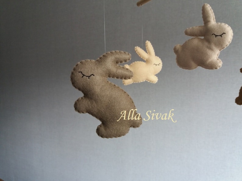 May include: A mobile with three felt bunny toys hanging from strings. The bunnies are brown and beige with black eyes and are in various poses. The mobile is hanging from a ceiling.