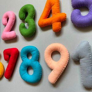 May include: Felt numbers 1 to 9 in various colours, including pink, green, orange, purple, red, yellow, blue, and grey. The numbers are stitched together with white thread.