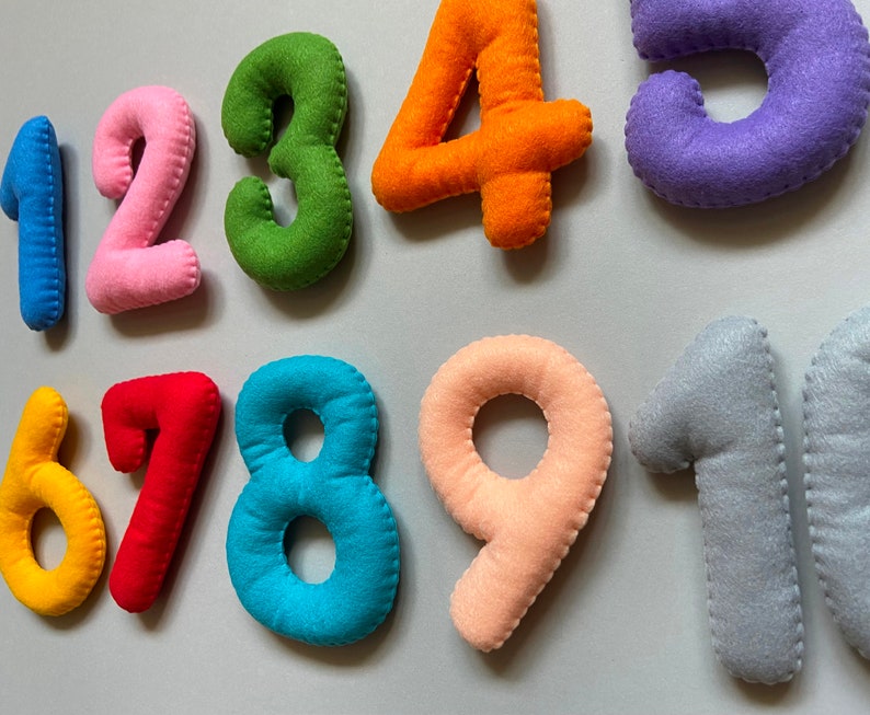 May include: Felt numbers 1 to 9 in various colours, including pink, green, orange, purple, red, yellow, blue, and grey. The numbers are stitched together with white thread.