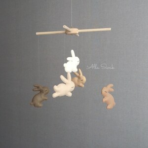 May include: A wooden mobile with five felt bunny toys hanging from it. The bunnies are in shades of brown and white. The mobile is hanging from a wooden rod.
