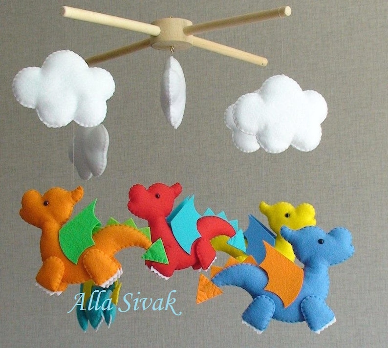 May include: A baby mobile with four felt dragons in orange, red, blue, and yellow with green, blue, and orange wings. The dragons are hanging from a wooden frame with three white felt clouds.