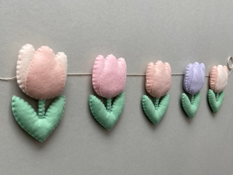 May include: A garland of felt tulips in pink, light purple, and green. The garland is strung on a white thread and would be a cute addition to a nursery or child's room.