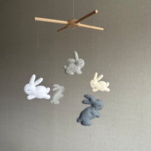May include: A wooden baby mobile with five felt bunny toys. The bunnies are in shades of gray, white, and beige. The mobile is suspended from a wooden crossbar.