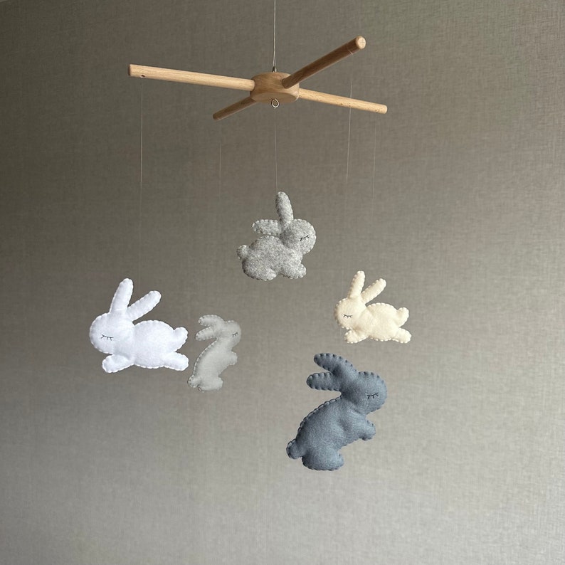 May include: A wooden baby mobile with five felt bunny toys. The bunnies are in shades of gray, white, and beige. The mobile is suspended from a wooden crossbar.