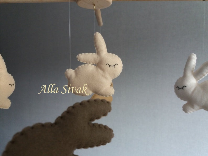 May include: A mobile with three felt bunny toys. Two bunnies are white with black eyes and the third bunny is brown with black eyes. The mobile is hanging from a wooden dowel.