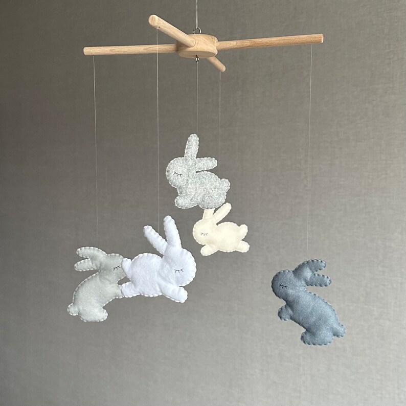 May include: A wooden baby mobile with four felt bunny toys hanging from it. The bunnies are in shades of gray, white, and cream.
