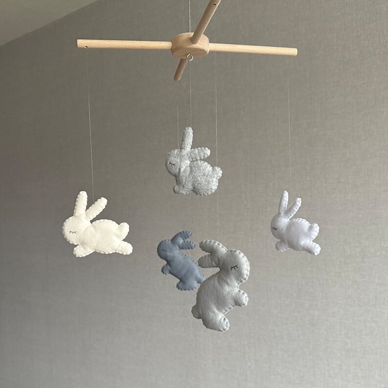 May include: A wooden mobile with five felt bunny toys. The bunnies are in shades of gray and white. The mobile is suspended from a wooden crossbar.
