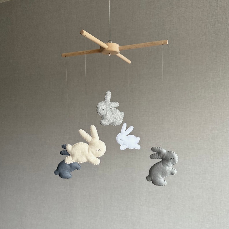 May include: A wooden mobile with four felt bunny toys hanging from it. The bunnies are in shades of gray and white.