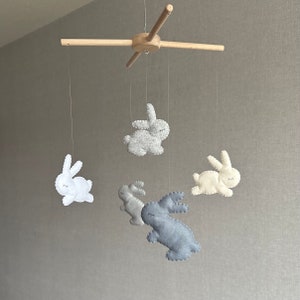 May include: A wooden mobile with five felt bunny toys hanging from it. The bunnies are in shades of gray, white, and light blue.