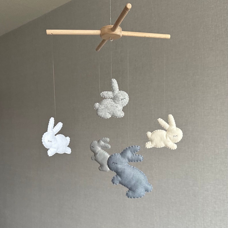 May include: A wooden mobile with five felt bunny toys hanging from it. The bunnies are in shades of gray, white, and light blue.