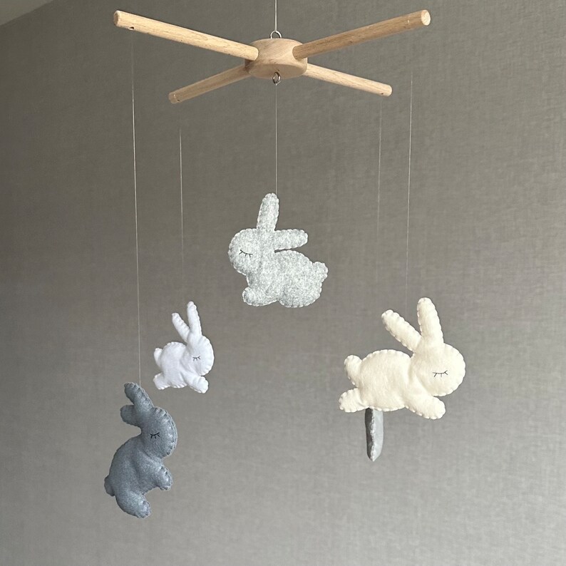 May include: A wooden mobile with four felt bunny toys. The bunnies are in shades of gray and white. The mobile is suspended from a wooden crossbar.