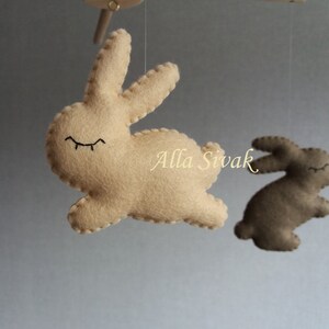 May include: A beige felt bunny with closed eyes hangs from a mobile. A second brown felt bunny is visible in the background.