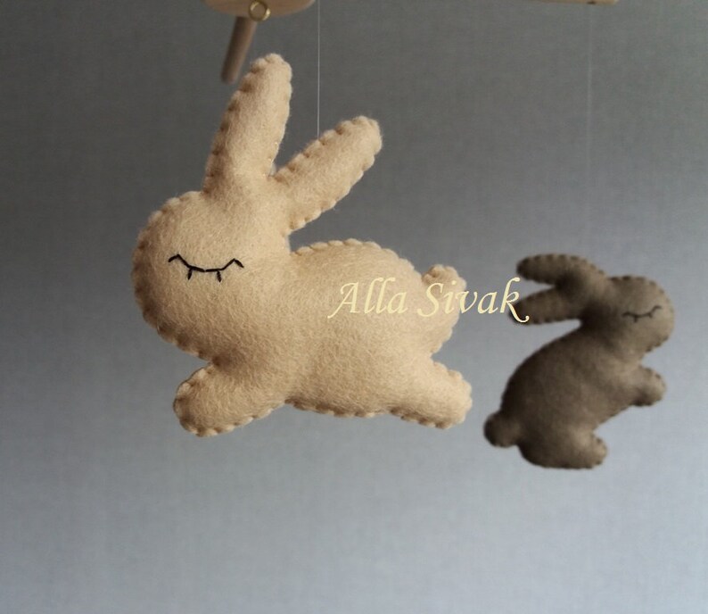 May include: A beige felt bunny with closed eyes hangs from a mobile. A second brown felt bunny is visible in the background.