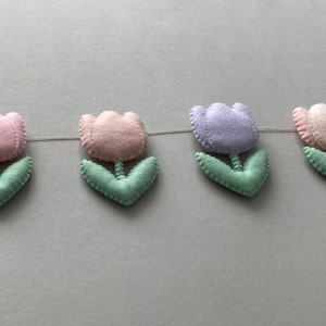 May include: A garland of felt tulips in pink, purple, and white with green leaves. The garland is strung together with white thread.