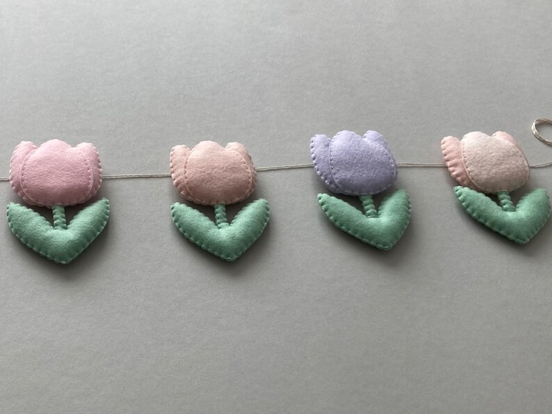 May include: A garland of felt tulips in pink, purple, and white with green leaves. The garland is strung together with white thread.
